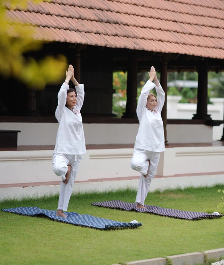 Two guests in white practising a tree pose on mats in front of a traditional building